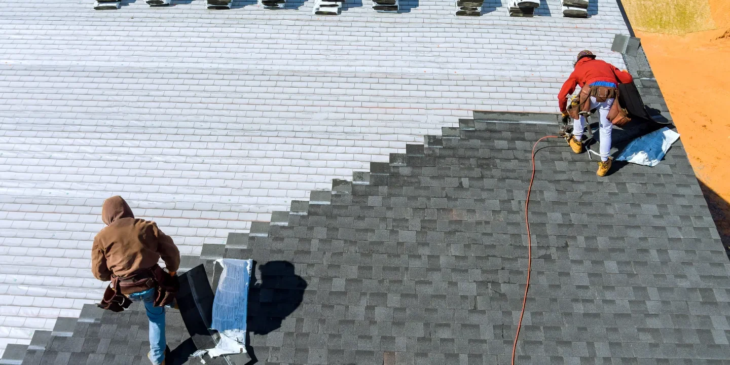 Two professional roofers are seen installing a roof on a house, with one roofer securing shingles and the other managing roofing materials during the roof installation process.